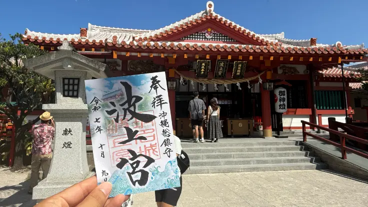波上宮の拝殿を背景に、御朱印を手に記念撮影をする参拝客。観光客にも人気の御朱印スポットになっている。