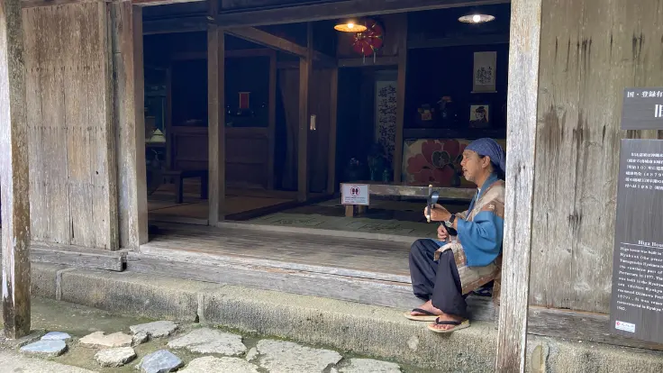 琉球村の古民家で三線を弾きながら沖縄の歌を披露するおじぃ。赤瓦屋根の下で穏やかな時間が流れる。