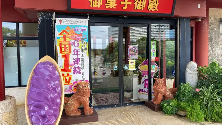お菓子御殿 読谷本店の正面入口。紅いもカラーの建物が印象的で、観光客が立ち寄る姿も見える。