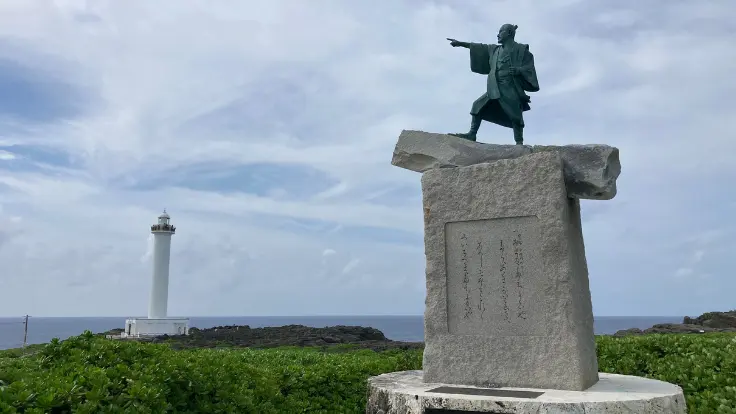残波岬に立つ泰期像と、その背後にそびえる白い灯台。空と海が広がる絶景の中にたたずんでいる。
