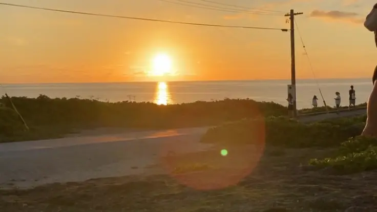 残波岬の灯台下で、夕日に照らされる人影。海に沈む太陽が空と海を黄金色に染めている。